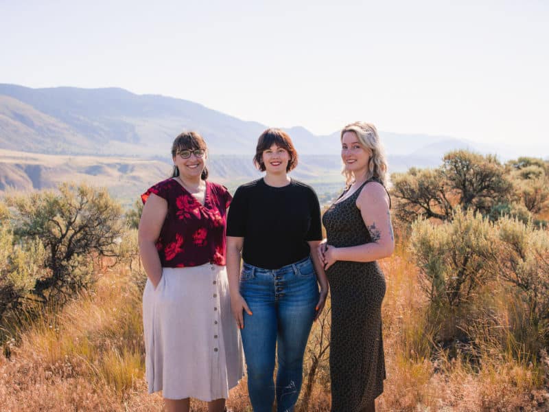 Lyssa Martin, Brandi Schier and Kyra Grubb stand together in a brush area in the Kamloops region. It's a sunny summer day. Everyone is smiling in the direction of the camera.