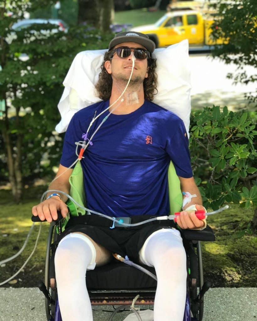 A person is shown in a hospital wheelchair with sunglasses on and tubes in his mouth.
