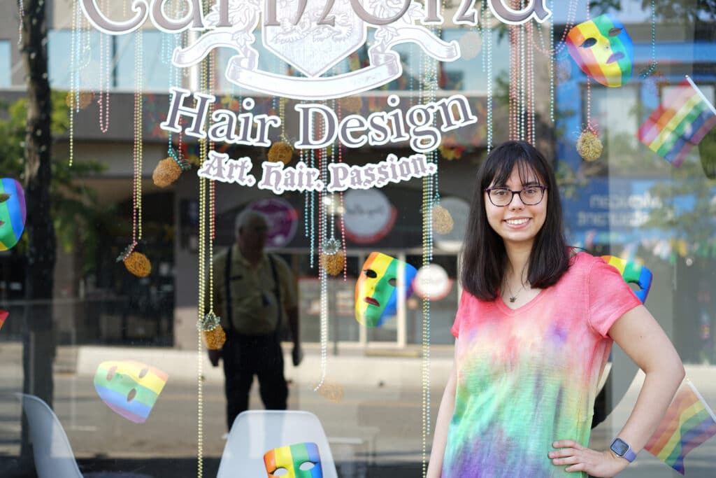 A person with long brown hair stands in front of a rainbow coloured window display smiling in a rainbow tie die shirt.