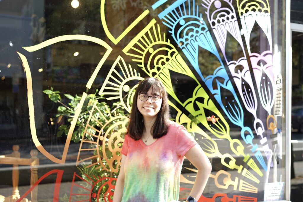 A person with long brown hair stands in front of a yellow blue and white mosaic painting, smiling.