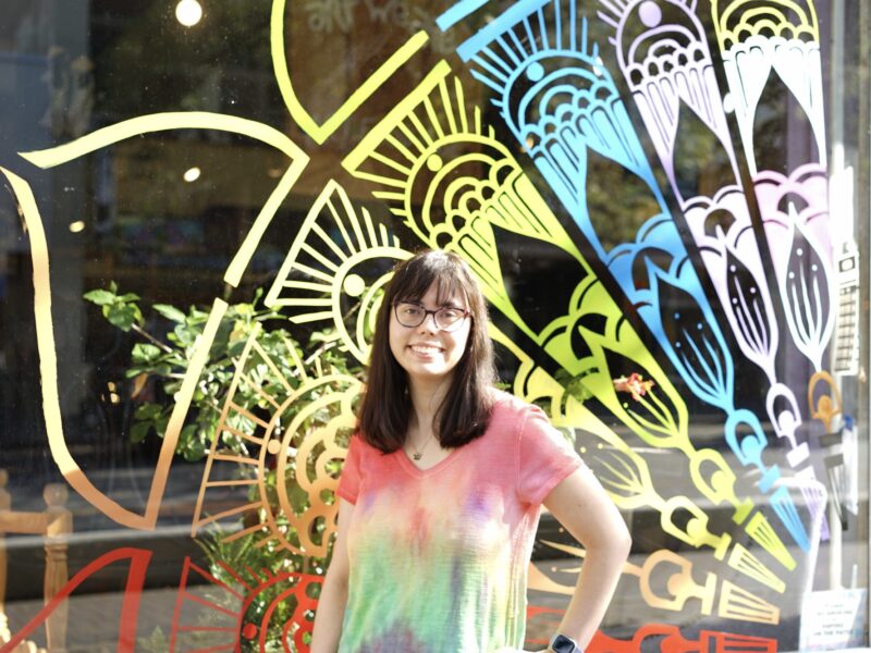 A person with long brown hair stands in front of a yellow blue and white mosaic painting, smiling.