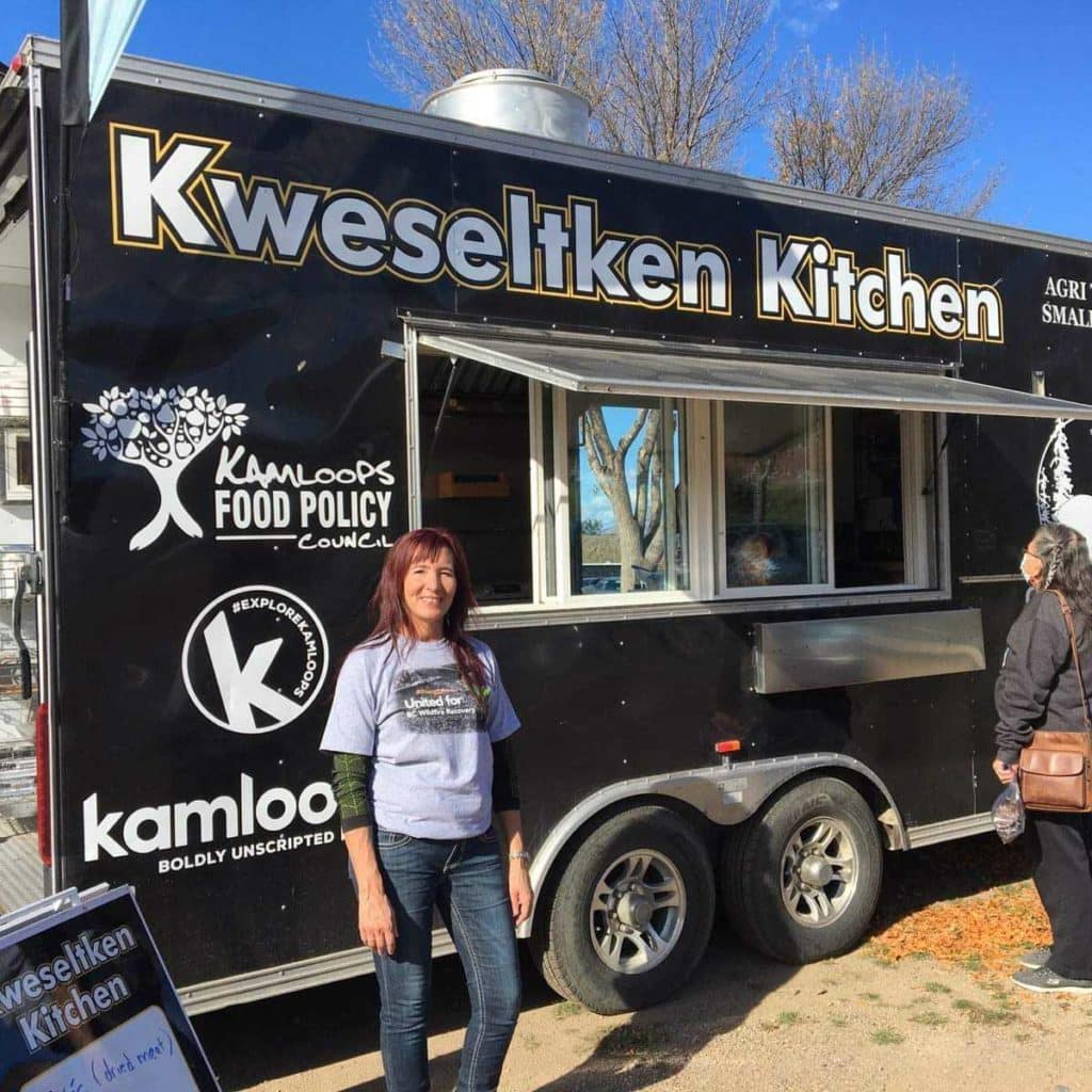 A woman stands in front of a black mobile kitchen truck with the words Kweselken Kitchen.