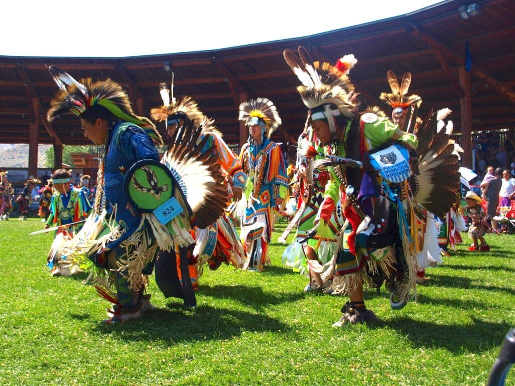 Dancers display their feathered and beded regalia through movement on the grass surrounded by stadium seating outdoors. during Saturday's Grand Entry at the 41st annual Kamloopa Powwow. 
