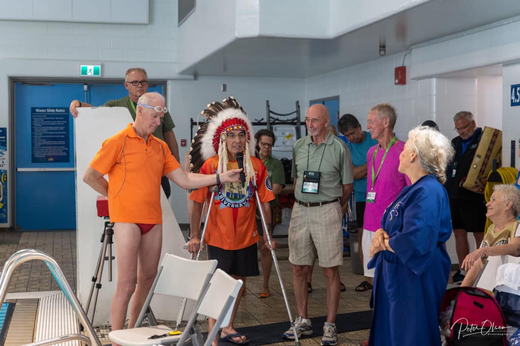 A person in an orange shirt and headdress makes a speech surrounded by people beside an indoor pool.