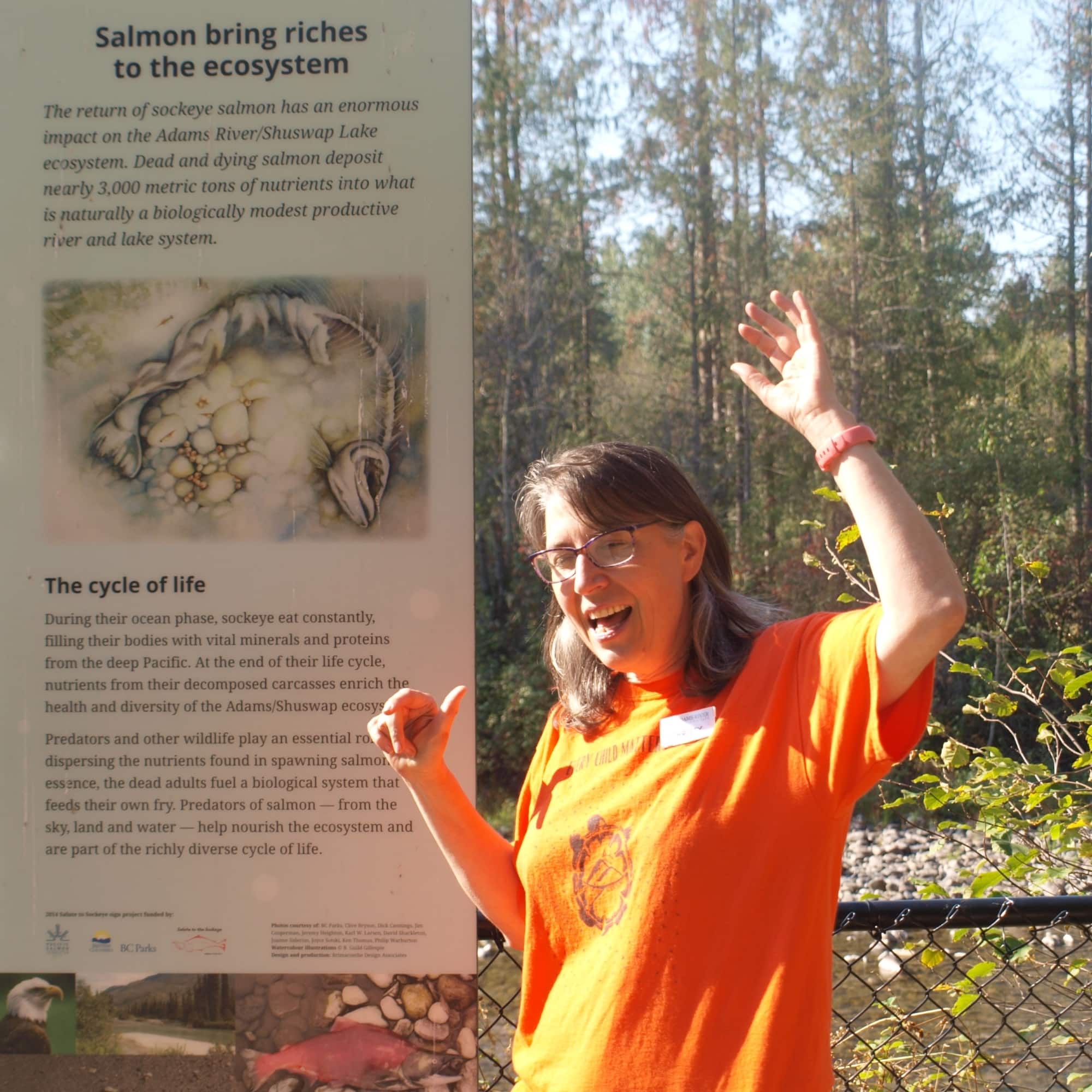 Christy Wright, wearing orange, stands in front of the river. Next to her is a sign with information about sockeye salmon, their contribution to the river and their cycle of life.