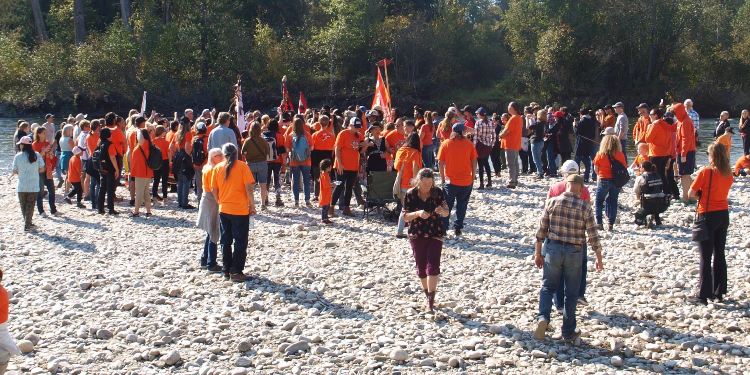 More gatherers can be seen on the river's pebble beach, most wearing orange. At the front of the crowd, different flags are held up.