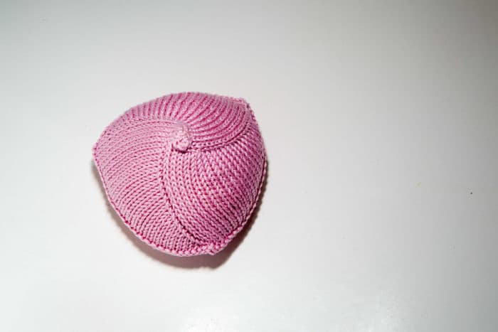 Knitted Knockers, a handmade breast prosthesis, sits against a white background. The prosthesis is made of bright pink yarn and shaped like a breast.