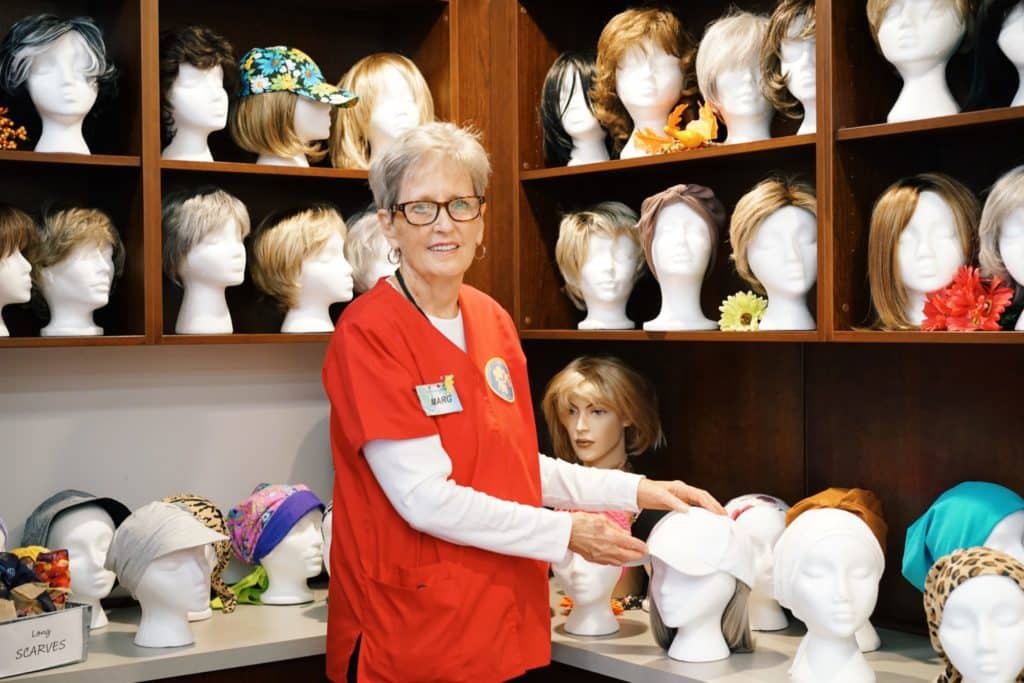 Marg Marshall stands dressed in a bright red shirt in front of the library of wigs. A variety of wigs in different cuts and colours surround her as well as caps and hats in an array of styles.
