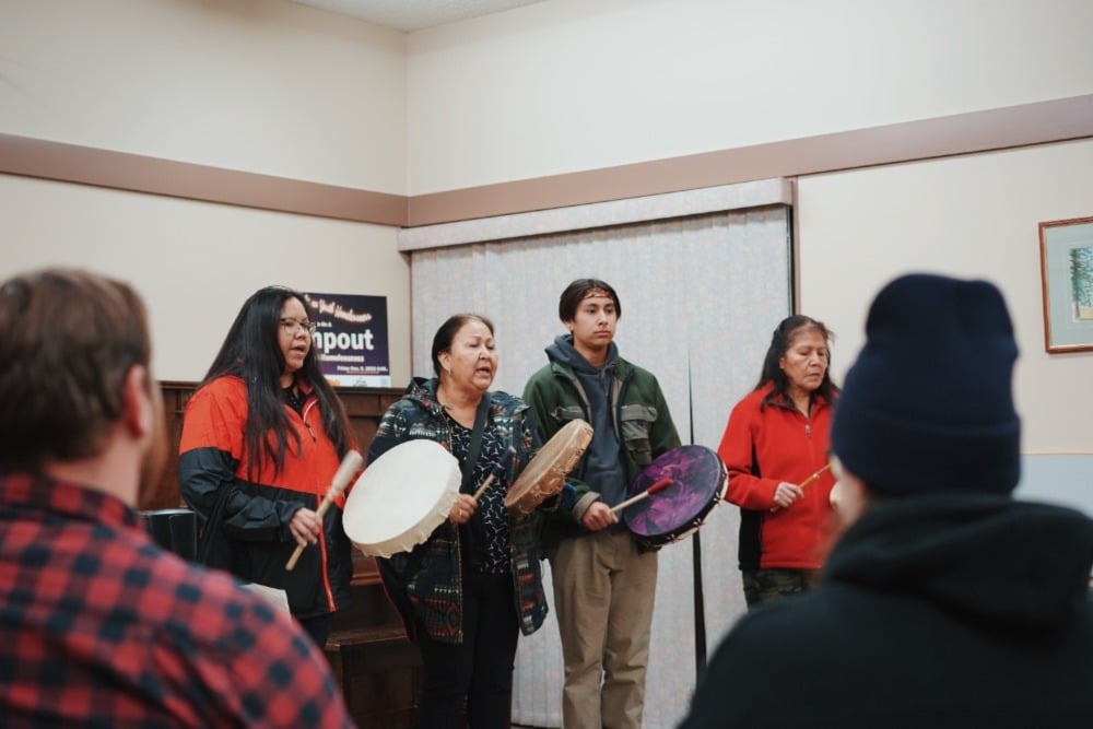 Four members of the Kamloops Aboriginal Friendship Centre sing and drum at the front of a room, while attendees watch.
