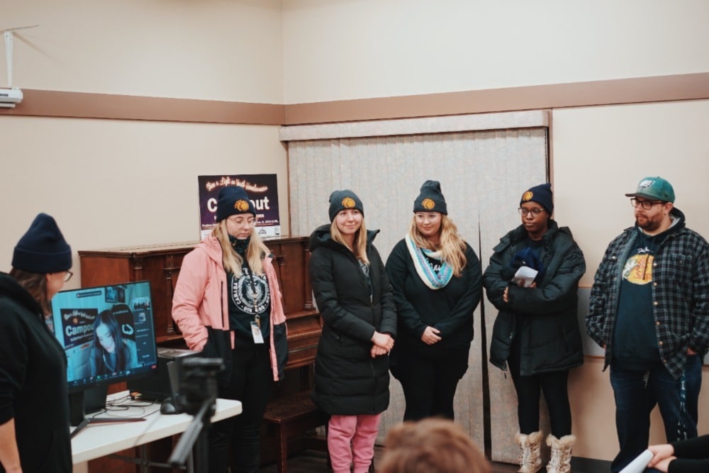 Six individuals wearing winter jackets and A Way Home Kamloops toques stand at the front of a room. Another person is seen presenting on a video call shown on a computer monitor.