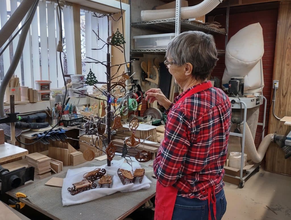 Hélène, wearing a red flannel shirt and a red apron, hangs ornaments on a tree-shaped display. She stands in her woodworking workshop. Proceeds from the ornaments will benefit Kamloops charities.