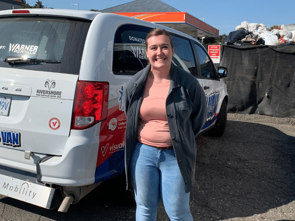 Sydney Mattis stands smiling in front of a large silver van with different logos for local charities and businesses on it.