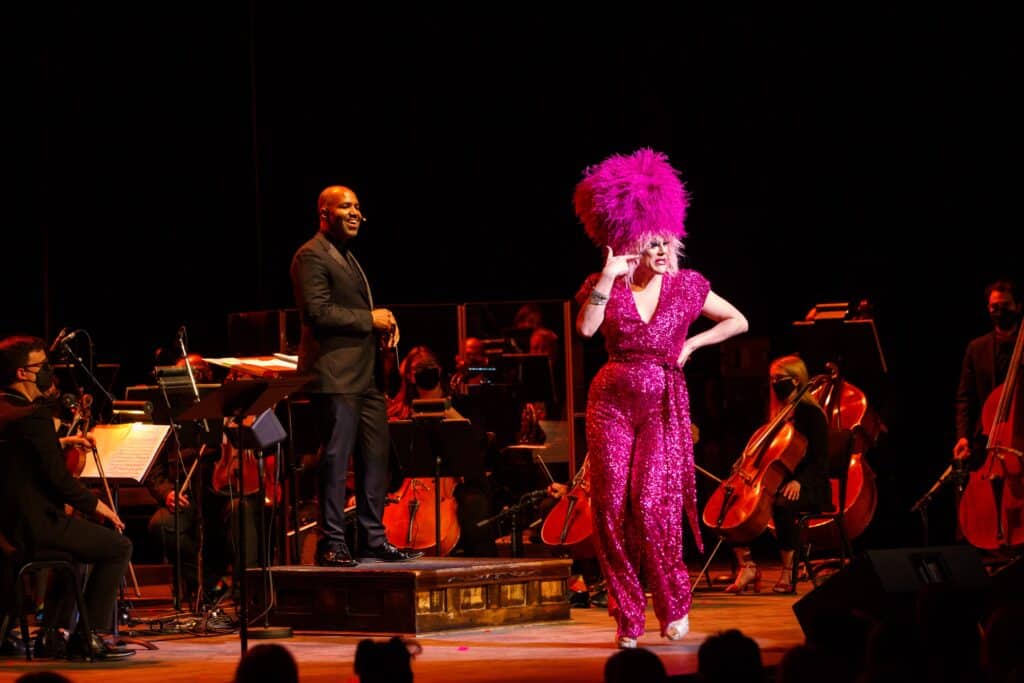 Thorgy Thor, a multi-instrumentalist and comedic drag performer, performs alongside the Regina Symphony Orchestra in 2022. Thorgy wears a pink sequinned jumpsuit and a feathered pink wig. The symphony sits behind her in all black.