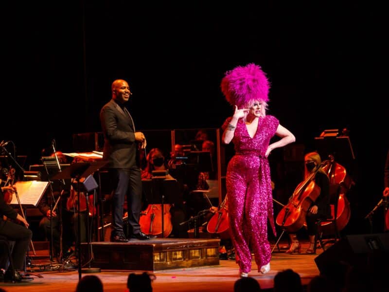 Thorgy Thor, a multi-instrumentalist and comedic drag performer, performs alongside the Regina Symphony Orchestra in 2022. Thorgy wears a pink sequinned jumpsuit and a feathered pink wig. The symphony sits behind her in all black.
