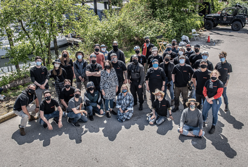 Around 30 people wearing masks and black clothing stand and sit together outside.