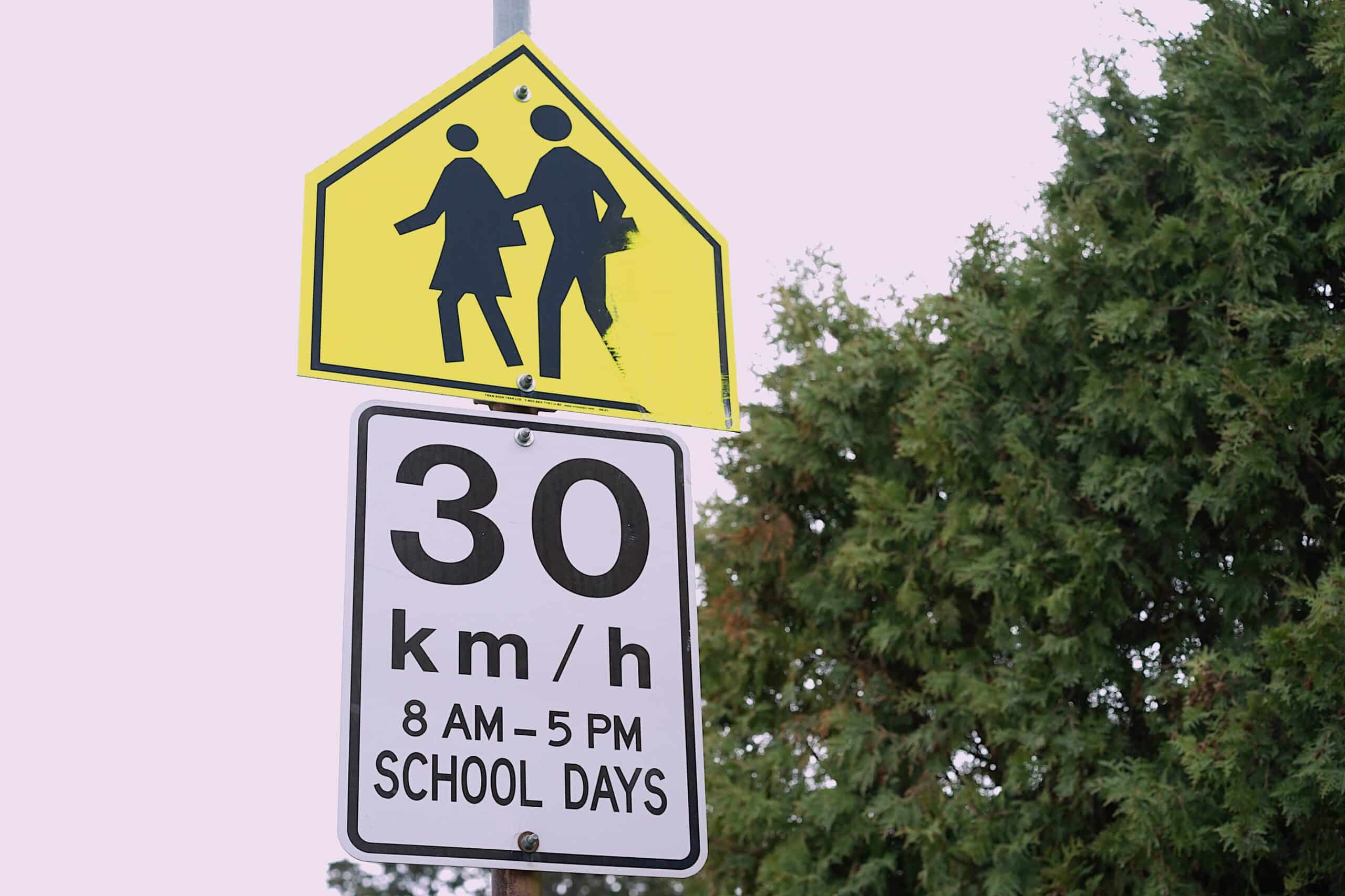 A school zone sign reads 30 km/ hr 8 AM - 5 PM SCHOOL DAYS as a safety precaution within a car-dependent culture