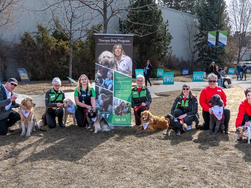 A group of people kneel outside with their dogs with a poster that reads Kamloops Therapy Dogs.