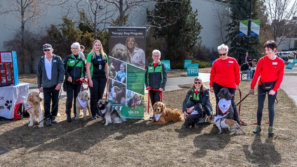 Seven people stand on a patch of grass. They are each wearing branded St. John Ambulance clothing. Each person also has a therapy dog on a leash with them. Each dog wears a St. John Ambulance bandana around their neck. Behind everyone is a sign that says "The St. John Ambulance Therapy Dog Program. Creating a memory of Joy in Your Community."