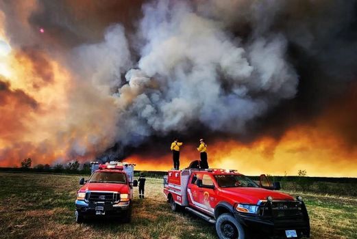 Image features large smoke plumes and flames from fires burning in northeastern B.C.. In the forefront, Kamloops Fire Rescue crew members stand on top of trucks parked a distance away from the fire.
