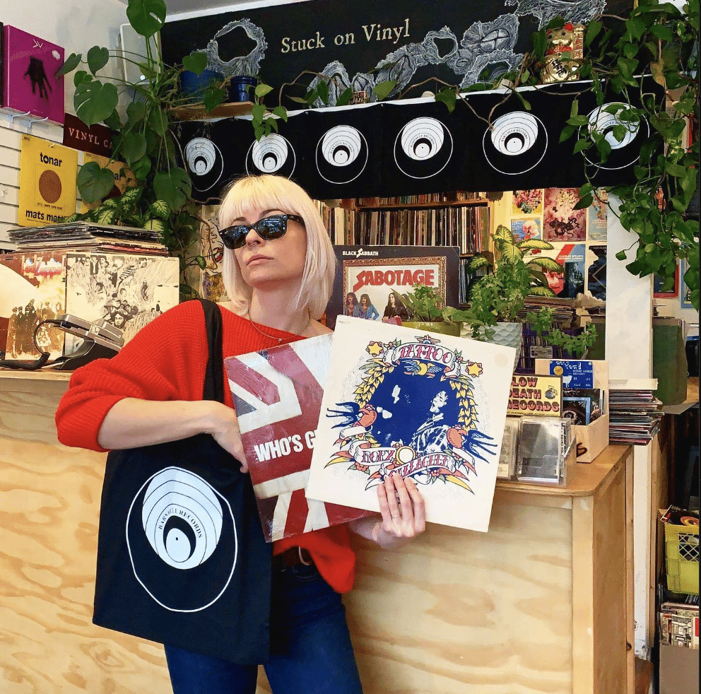 Jessie McGrath, co-owner of Barnacle Records, holds two records. She is wearing a red shirt and black sunglasses.