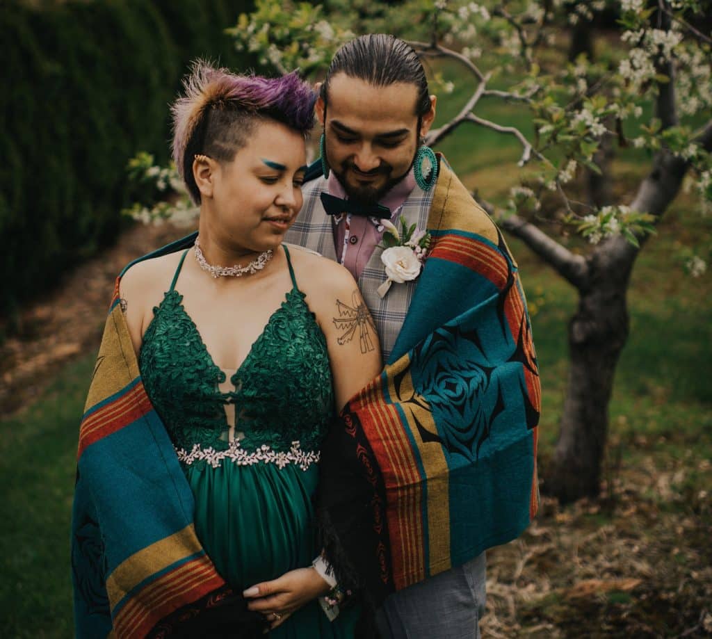  Joseph Jules-Peters (right) and Landon Sekwaw Peters (left)  embrace under a blanket. Landon wears a green dress and Joseph is in a plaid vest. There is a tree in the background, just out of focus. 