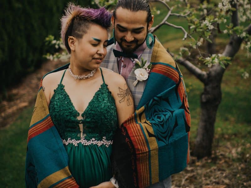 Joseph Jules-Peters (right) and Landon Sekwaw Peters (left) embrace under a blanket. Landon wears a green dress and Joseph is in a plaid vest. There is a tree in the background, just out of focus.