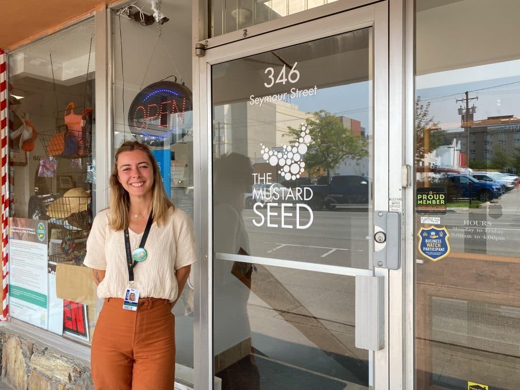 A woman wearing a white shirt and burnt orange pants stands in front of The Mustard Seed Thrift Store, where unhoused folk can come and shop for high heat essentials.
