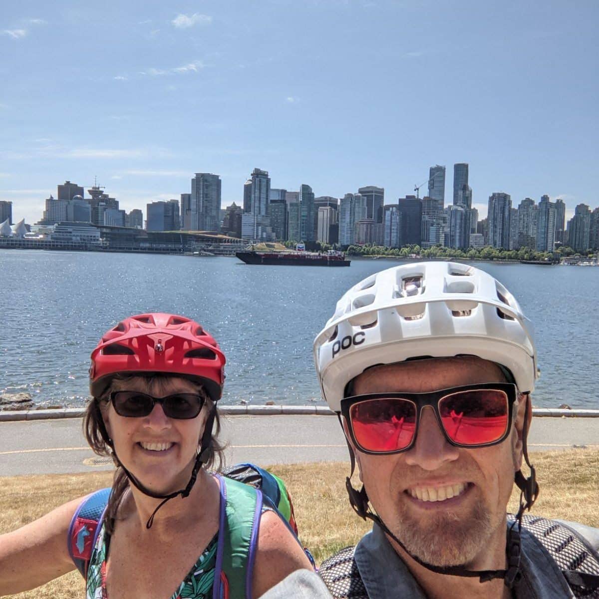 Two people stand with a city in the background. It is clear they have been biking as they are wearing helmets. The man in the photo is Lyall Alore and he is wearing a white helmet and black sunglasses. The woman in the photo is Deb Alore and she is wearing a red helmet with black sunglasses. They are both members of Stolen Bikes Kamloops.