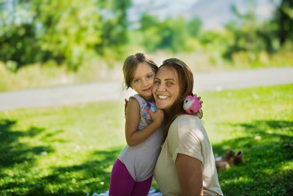 A girl hugs her mother around the neck seated in a grassy park.