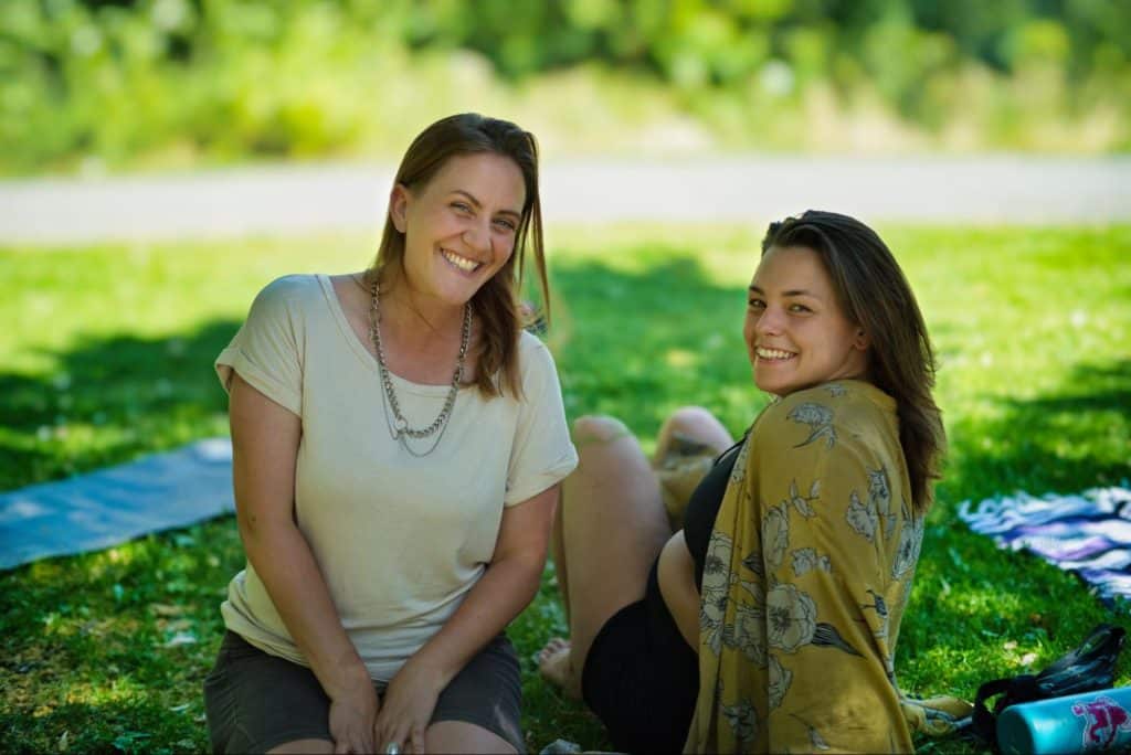 Two women sit and smile side by side in a grassy green park.