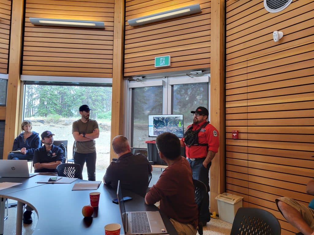 A group of people stand with serious faces in a room with screen displaying a map.