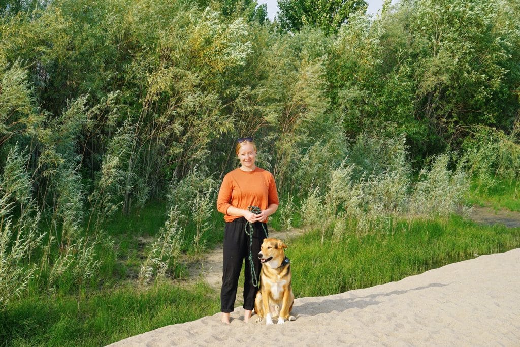 A woman stands to the left of her dog, a brown, black and white mixed breed. They are on sand, with vegetation in the background.