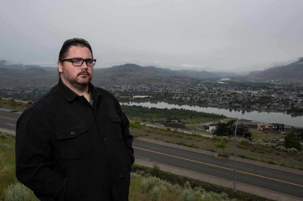 A man wearing black with black glasses and a beard stands with the mountains and river in the background. 