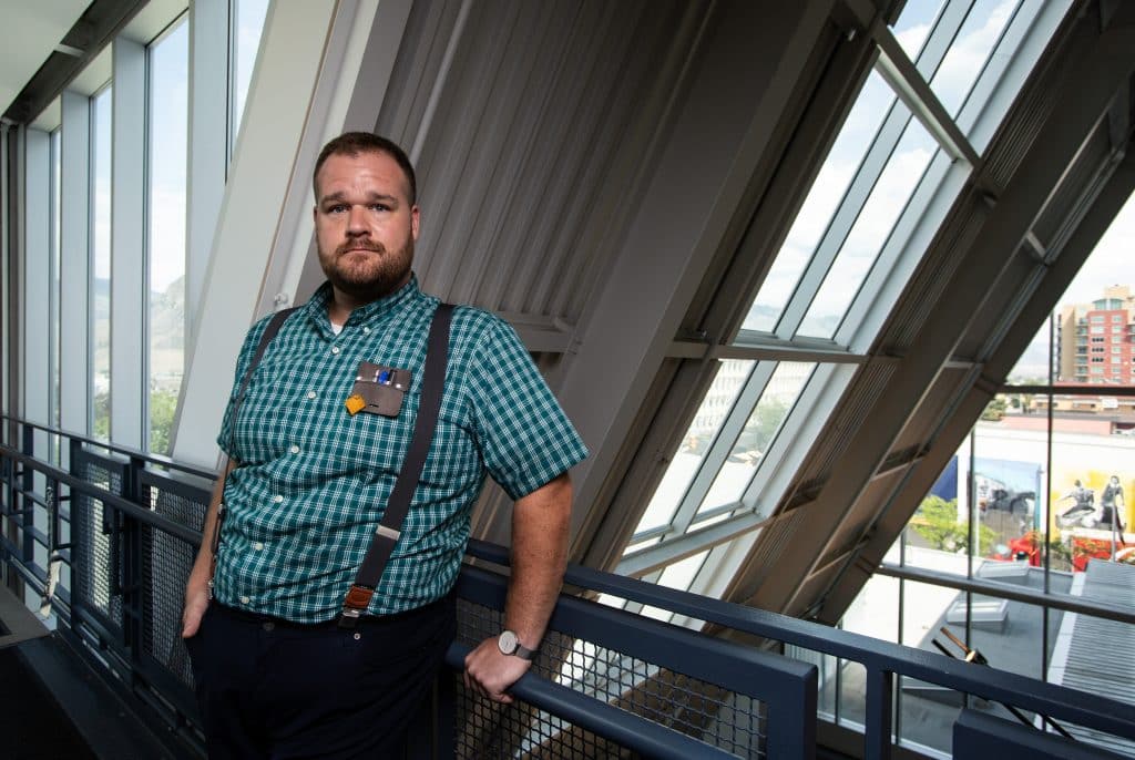 A person with suspenders and collared shirt stands next to an indoor balcony and window.