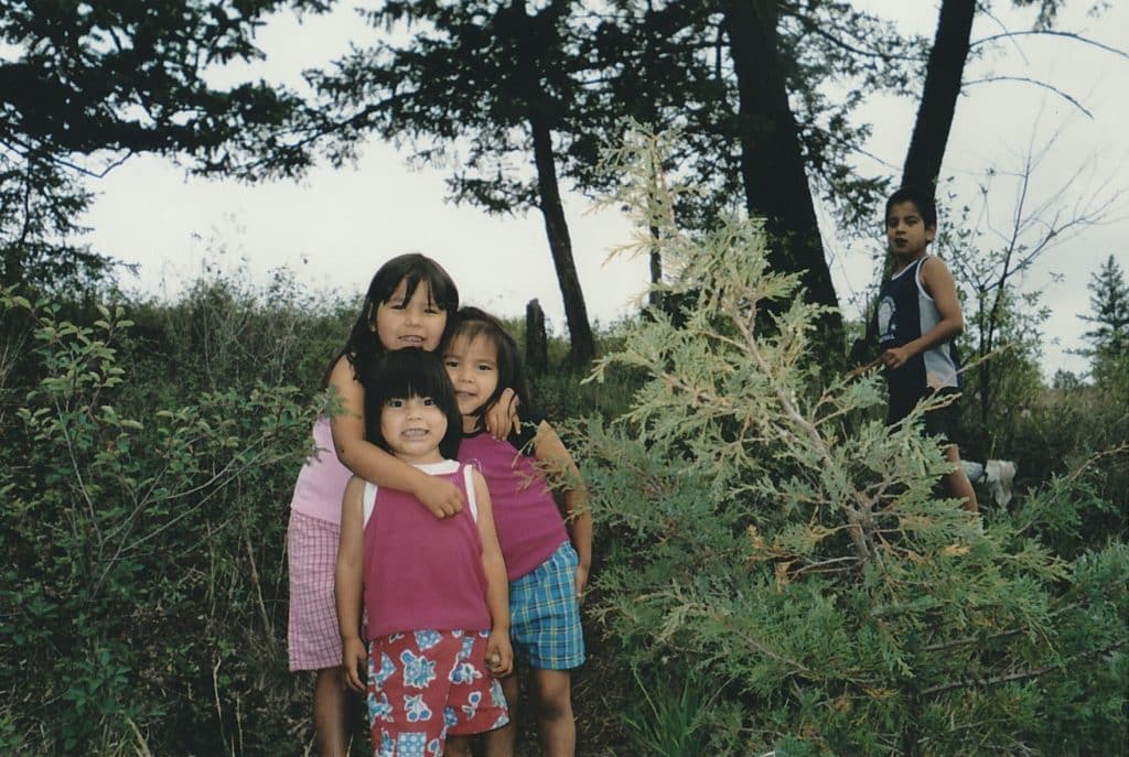 A taller child hugs two younger cousins in a forested area of Xats̓úll near Williams Lake.