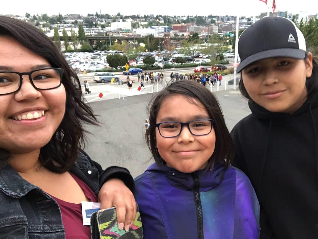A selfie shows a woman smiling with glasses and tow younger siblings beside her, also smiling.