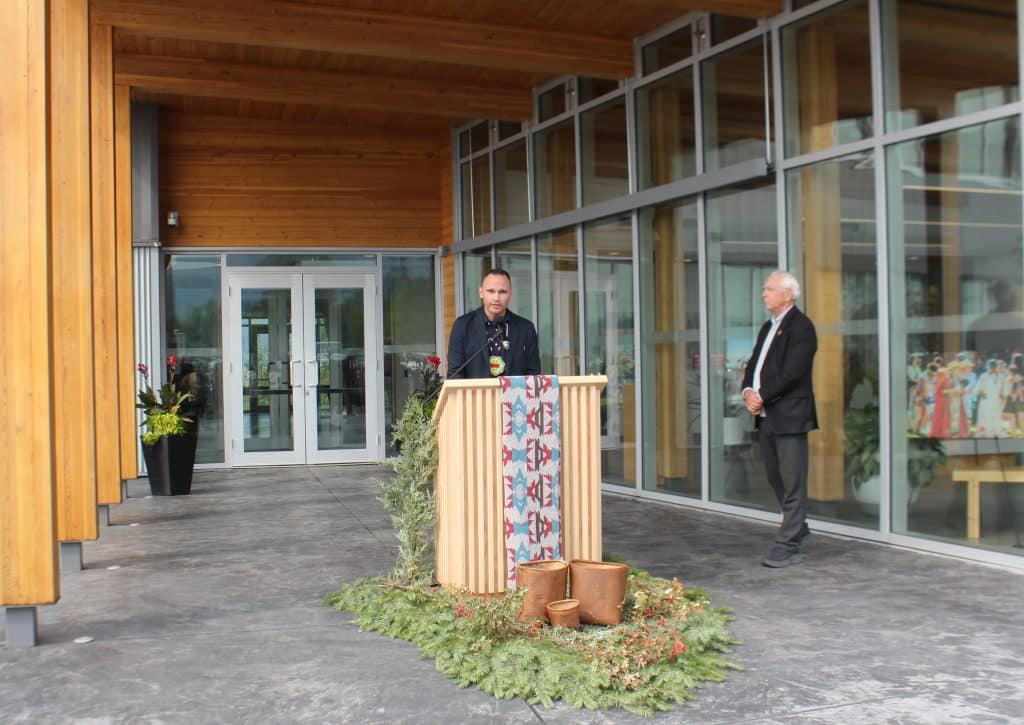 A man stands behind a podium with a mic, cedar boughs and a tapestry are draped from the front.