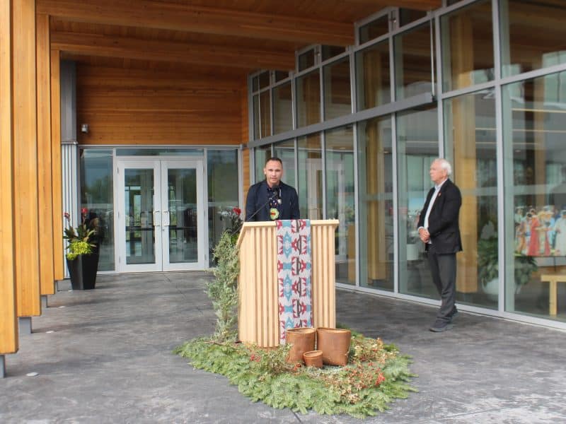 A man stands behind a podium with a mic, cedar boughs and a tapestry are draped from the front.