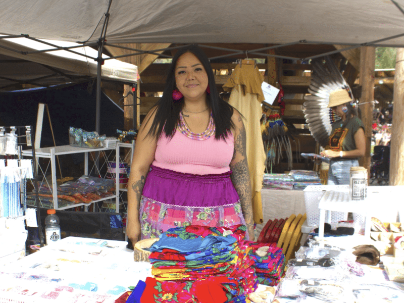 A woman smiles wearing a pink shirt, red earrings and a colourful ribbon skirt at a booth with bright fabrics and goods.