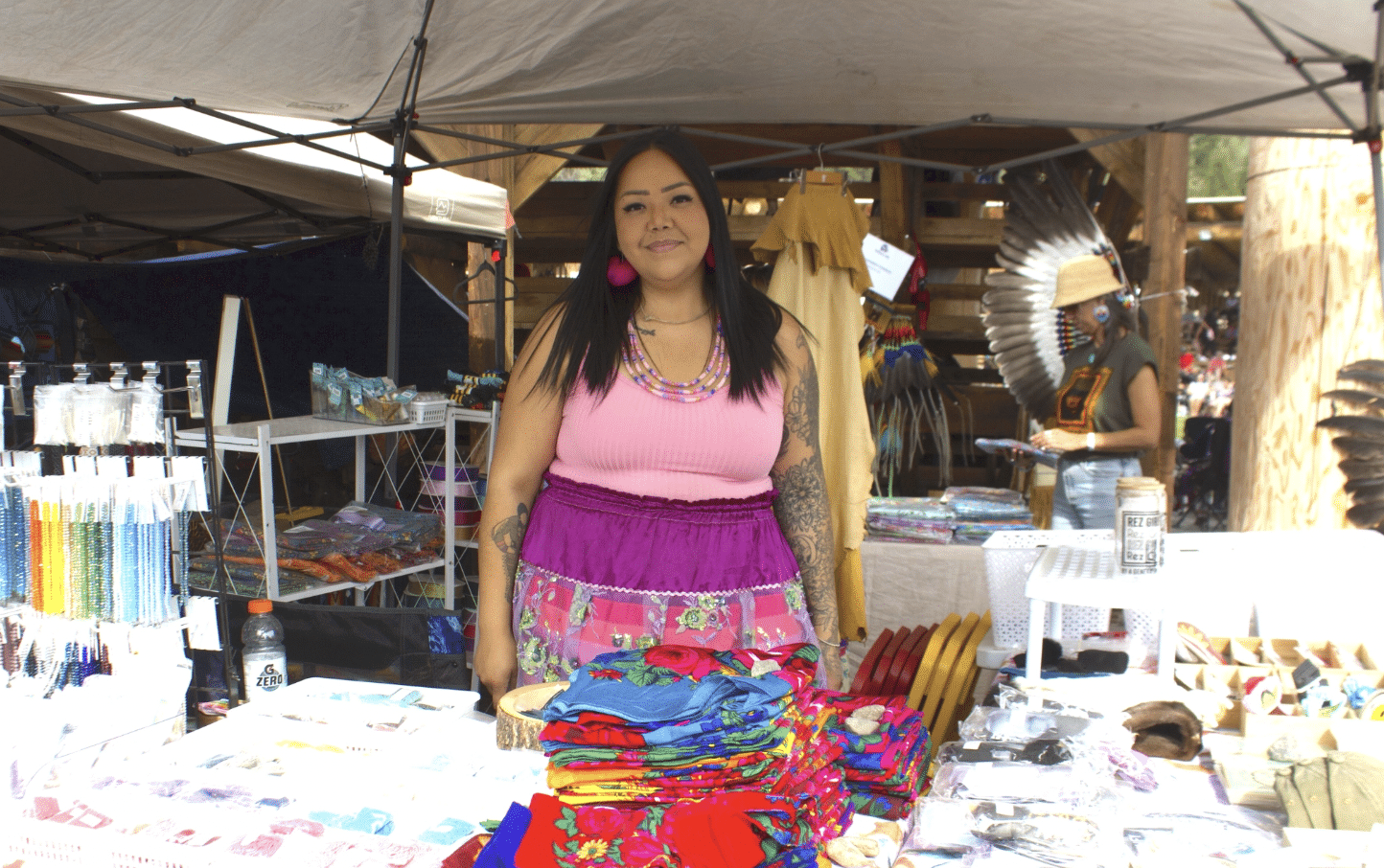 A woman smiles wearing a pink shirt, red earrings and a colourful ribbon skirt at a booth with bright fabrics and goods.