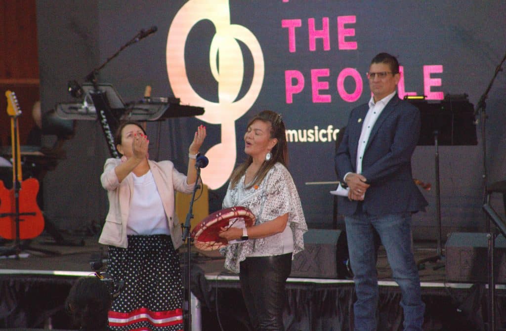 Kúkwpi7 Rosanne Casimir sings the Welcome Song before the performance. A screen reads "Music for the People" in the background and she holds a drum