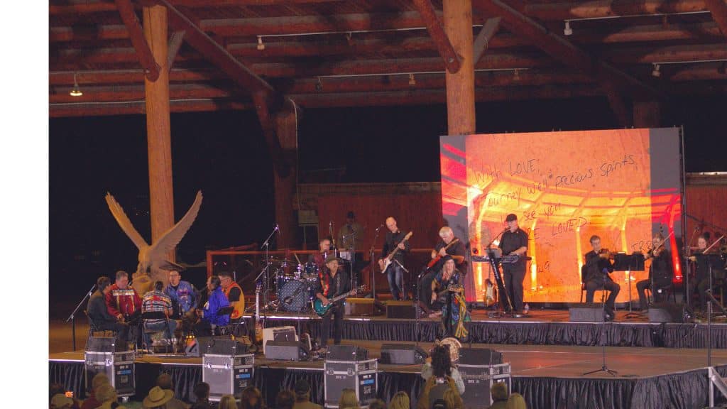 A bunch of artists with various regalia and instruments stand on stage with a bright orange screen in the background.
