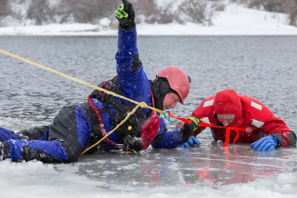 a member of Kamloops Search and Rescue in blue lays on ice, assisting someone in red.