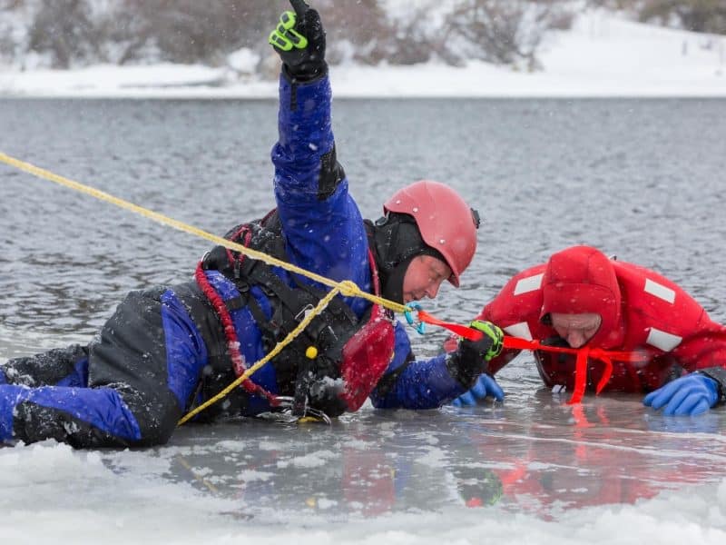 a member of Kamloops Search and Rescue in blue lays on ice, assisting someone in red.