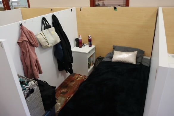 A cubby at Merit Place shows a single bed with a pillow, side table and clothes hanging on wall.