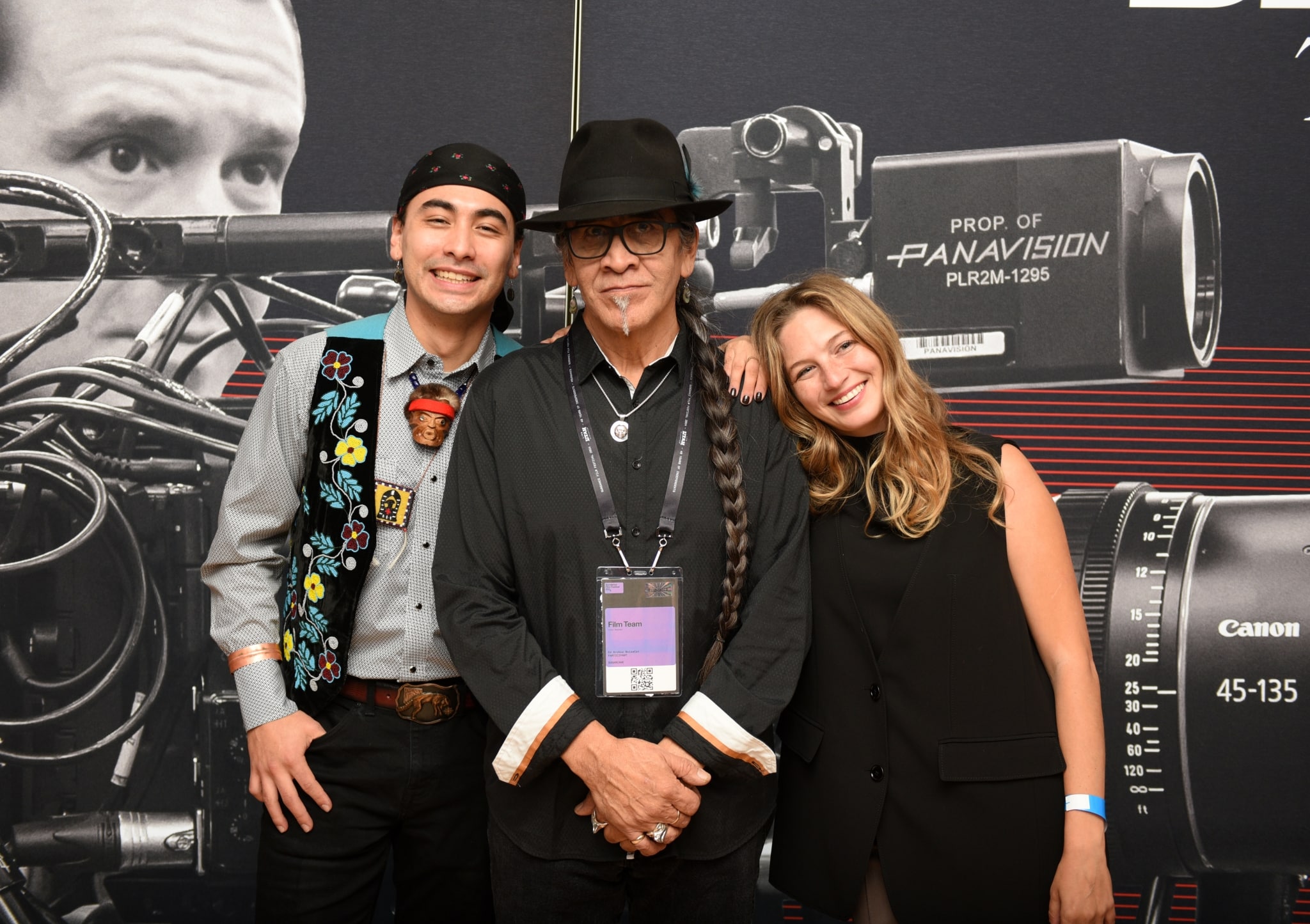 Three people stand amicably smiling. At centre is a man with a felt hat and long braid wearing a film press badge.
