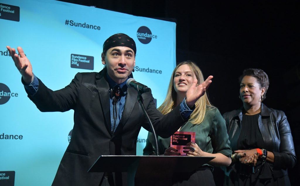 Julian Brave NoiseCat stands with open expressive arms at the podium for film award next to two women.