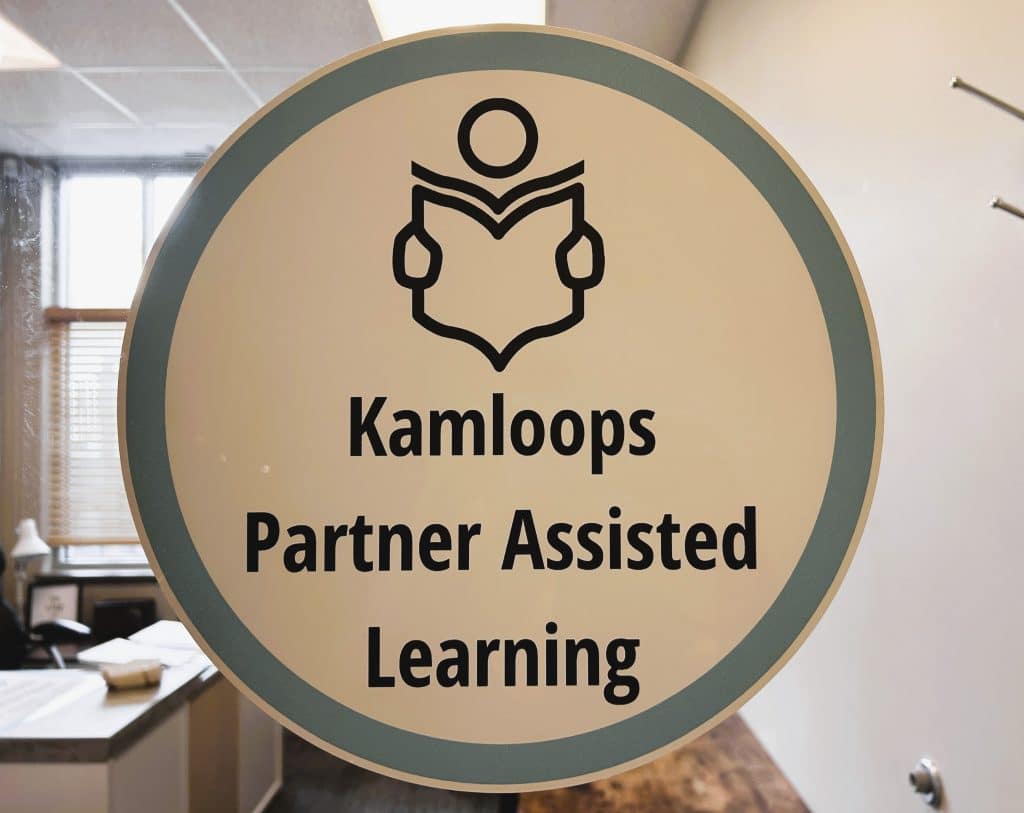 A photo of a sign with the Kamloops Partner Assisted Learning (KPAL) name and a small illustration of a figure reading a book.