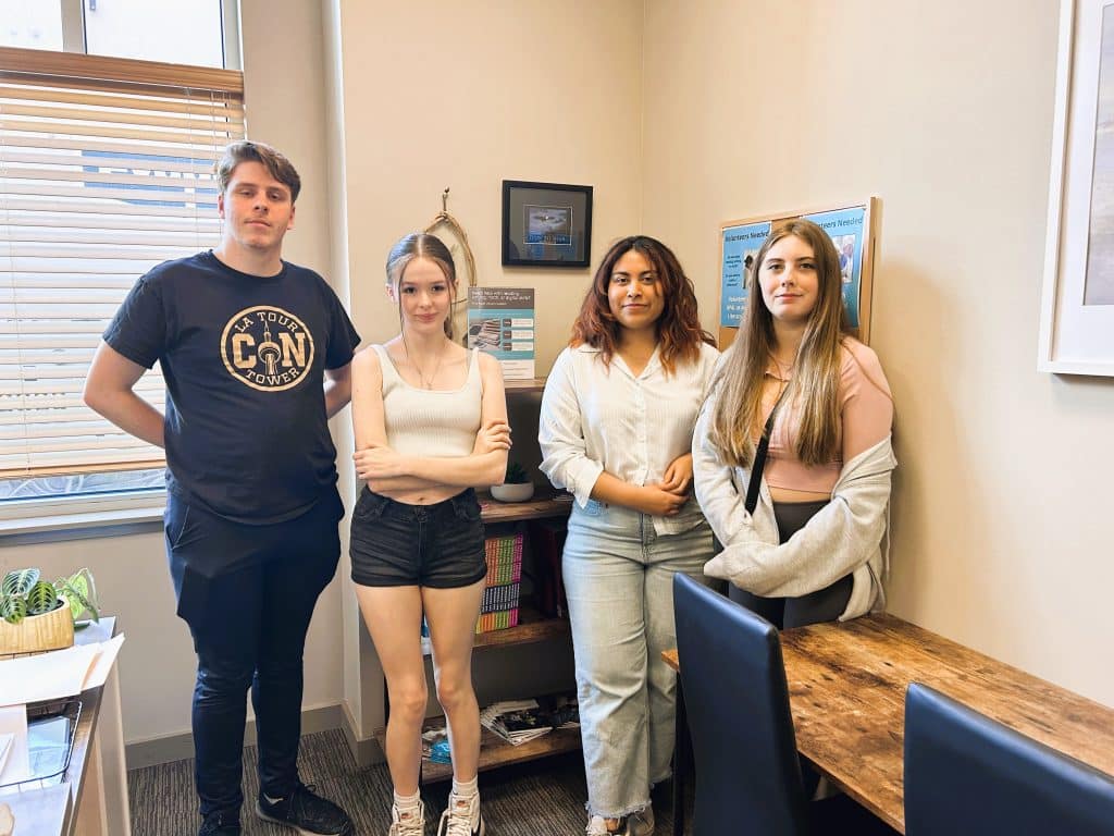 The photo shows four of the current youth volunteers standing side by side at the back of a room and looking towards the camera.