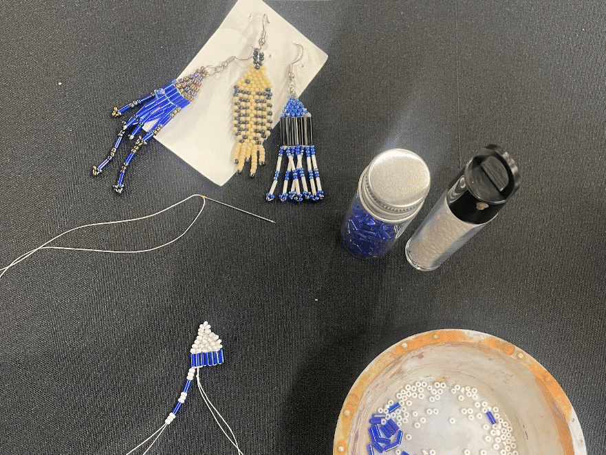 On top of a black table cloth lies the beaded earrings, two plastic vials of seed beads, a dish with seed beads, and a fourth beaded earring that is incomplete.